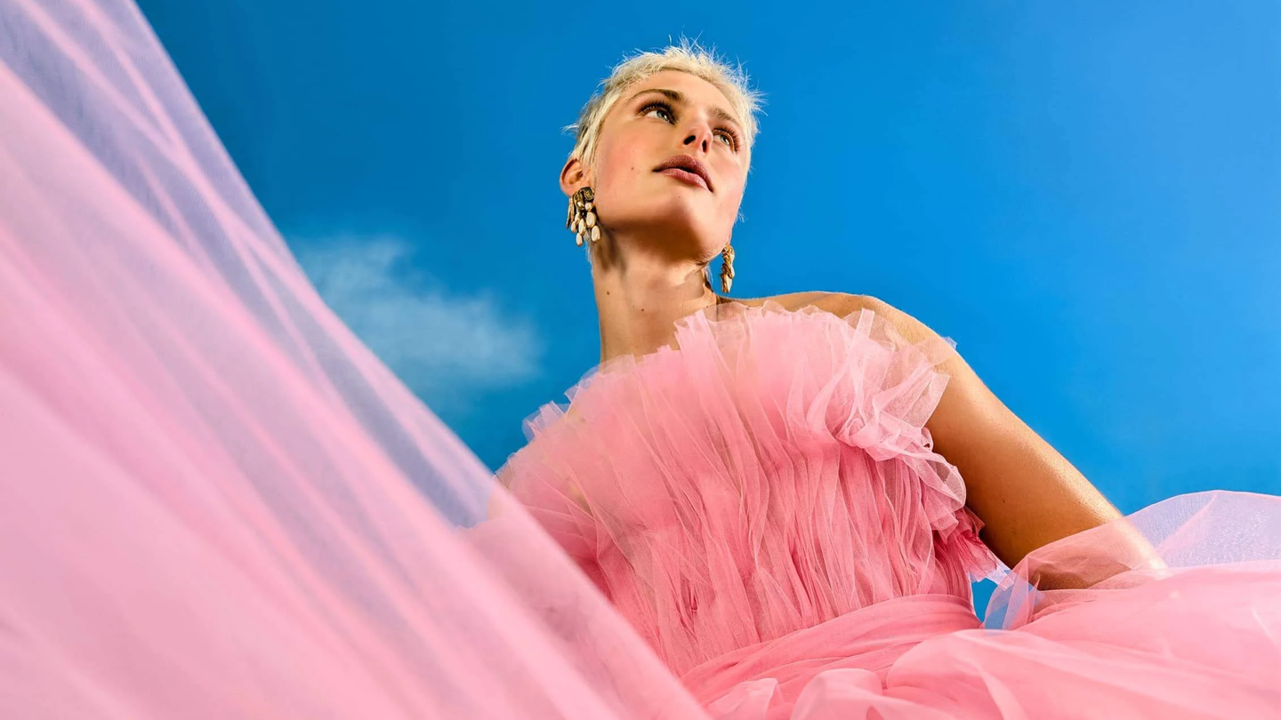 A fair-skinned person with short platinum-blond hair, pink lips, and dangling earrings wears a pink tulle dress.